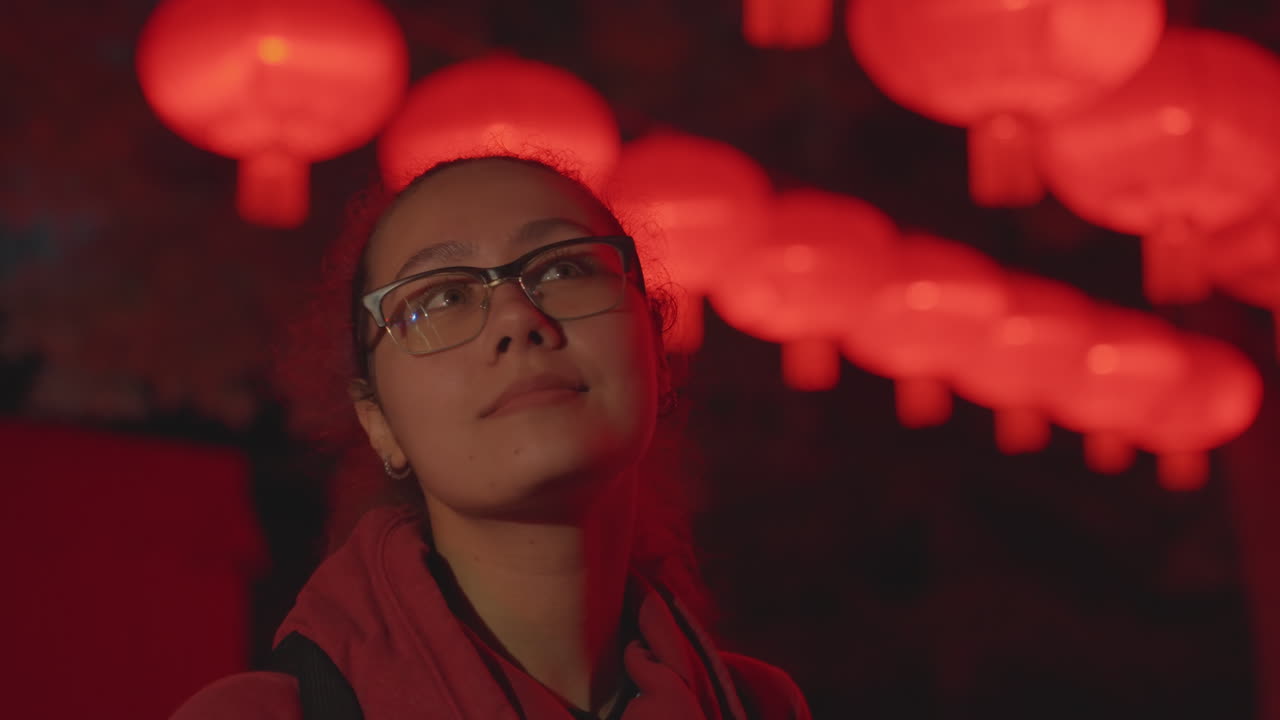 Mujer mirando linternas rojas en lo alto, con un suave reflejo en sus gafas y expresión tranquila en un primer plano de perfil con coleta, con una cálida luz roja de contorno y poca profundidad de campo, creando un ambiente de callejuela festiva tranquila.