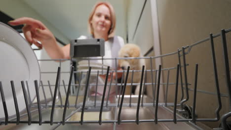 cute woman takes clean plates from the dishwasher. her puppy is next to her