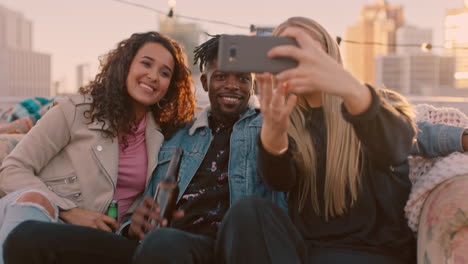 happy multi ethnic friends enjoying rooftop party drinking alcohol taking selfie photos using smartphone sharing fun weekend on social media at sunset