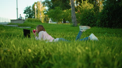 programmer lying on grass in park working with laptop while holding apple in hand, surrounded by lush greenery and trees with urban structures visible in blurred background under soft natural light