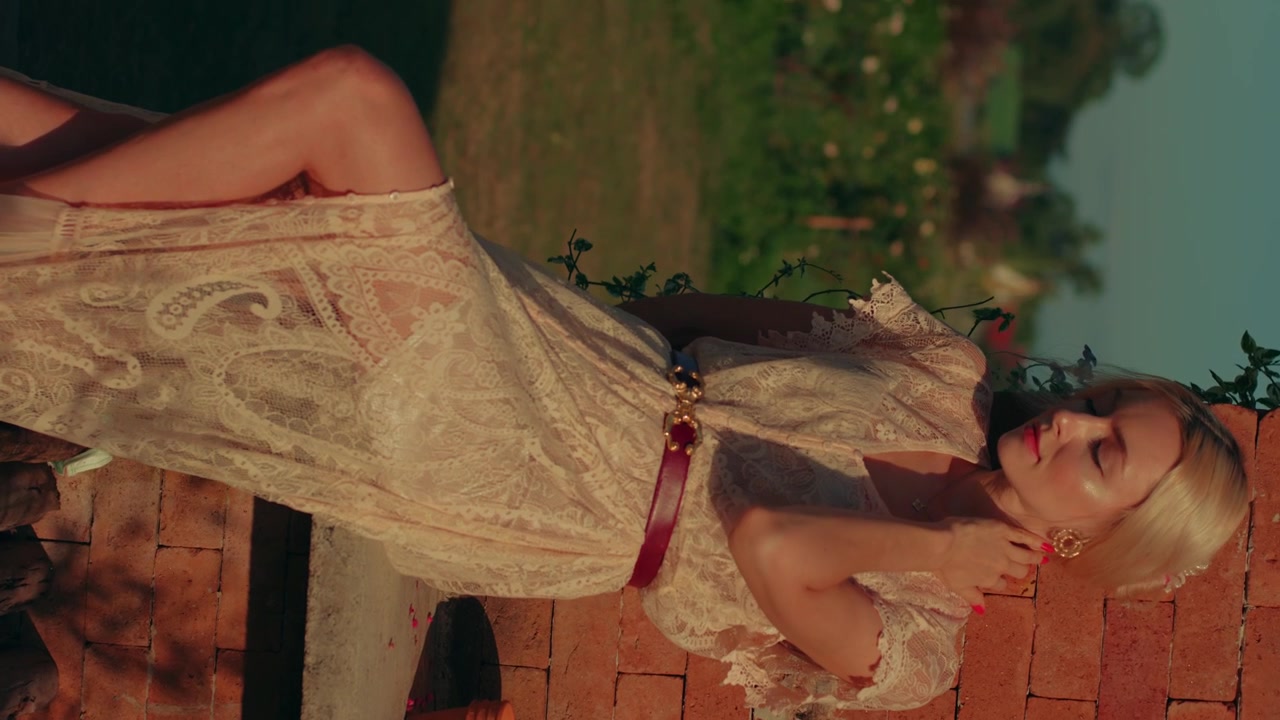 Donna elegante in abito di pizzo appoggiata a un muro di mattoni al tramonto.