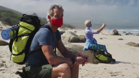 Senior-hiker-man-wearing-face-mask-with-backpacks-sitting-on-a-rock-and-senior-woman-taking-pictures