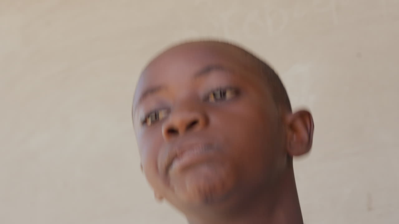 Menino negro de camisa azul rindo e dançando em close-up, com gestos expressivos de ombro e braço contra uma parede iluminada pelo sol, sorriso brilhante e ritmo animado, performance juvenil espontânea cheia de exuberância.