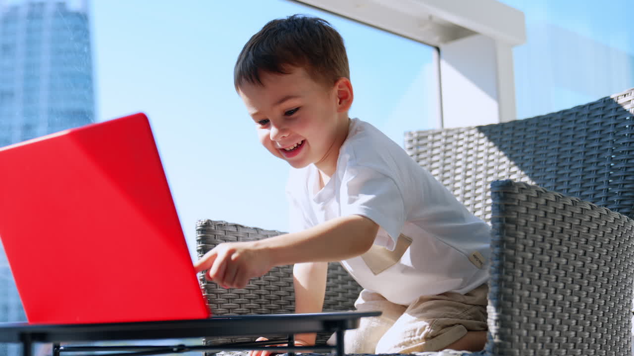 Boy happily plays on laptop. A cheerful boy sits in a bright space, focused on a red laptop while smiling. Sunlight pours through large windows