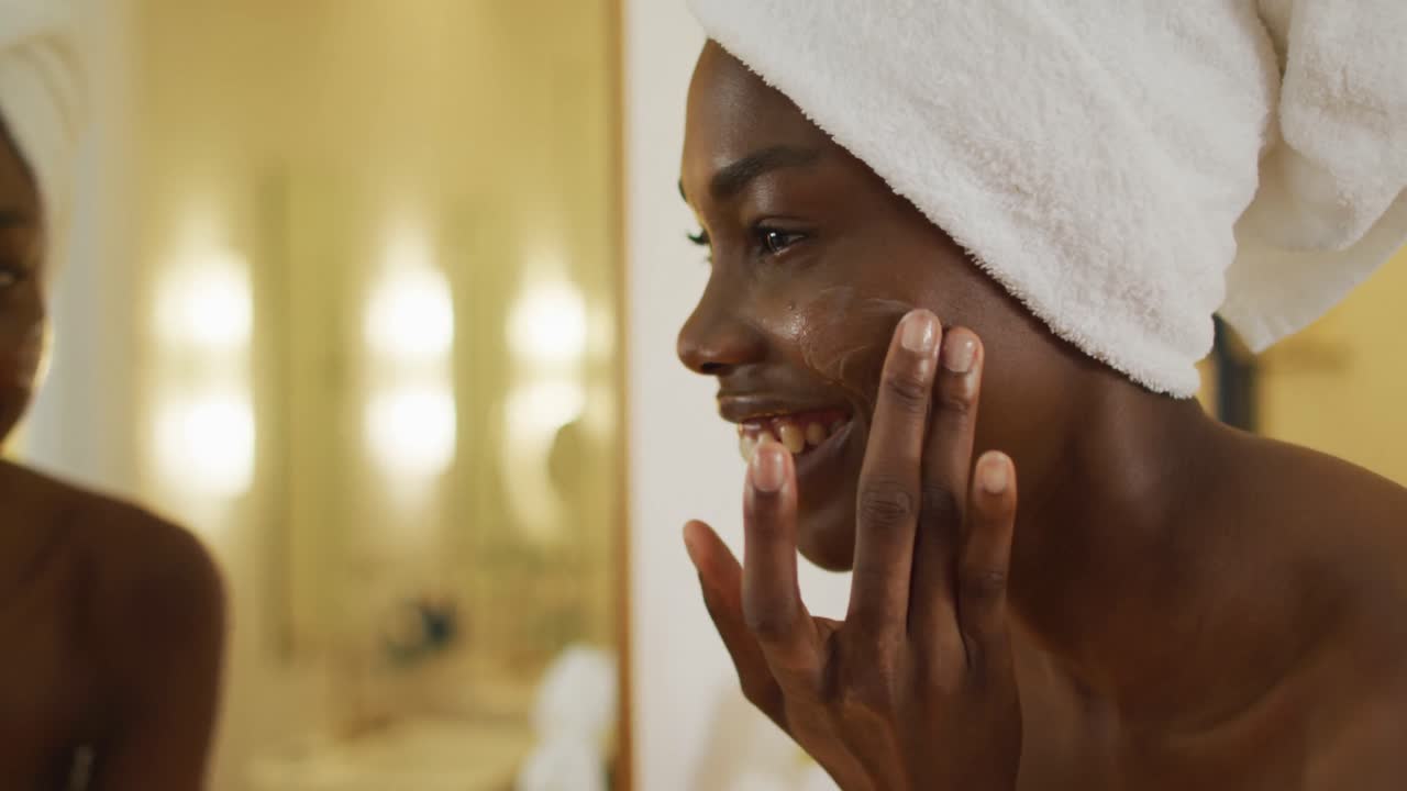 mujer afroamericana sonriente con una toalla mirándose en el espejo y usando crema en su cara en el baño