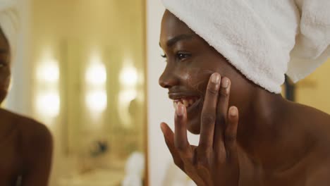 Lächelnde-Afroamerikanische-Frau-Mit-Handtuch,-Die-Im-Spiegel-Zuschaut-Und-Im-Badezimmer-Creme-Auf-Ihr-Gesicht-Aufträgt