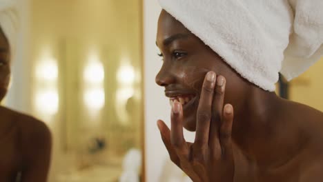 smiling african american woman with towel watching in mirror and using cream on her face in bathroom