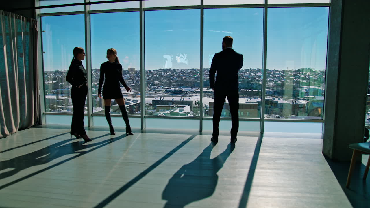 Young people indoors on large windows background. Beautiful business women talking about man. Businessman standing in office near windows in sunny day.