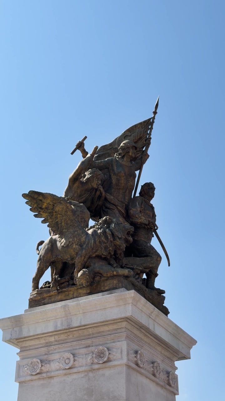 Monumento in bronzo con figure e un leone alato in cima a un piedistallo di pietra sotto un cielo sereno.
