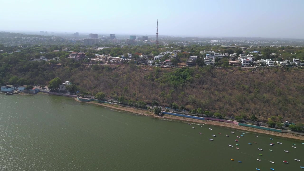 Filmato drone che si muove all'indietro sopra la riva del Lago Superiore, rivelando residenze collinari, pendii coperti da alberi, barche ancorate e viste panoramiche della città di Bhopal.