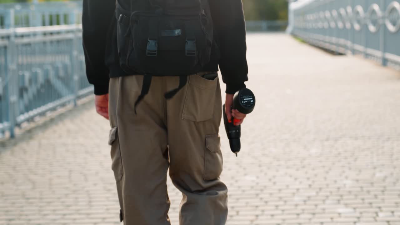 Uomo caucasico, fotografo di strada, cammina lungo un ponte con la macchina fotografica che pende dallo zaino. Indossa pantaloni cargo casual e abbigliamento a strati, in una scena di calda luce in controluce e solitudine urbana, con un'atmosfera narrativa da viaggio e creativa.