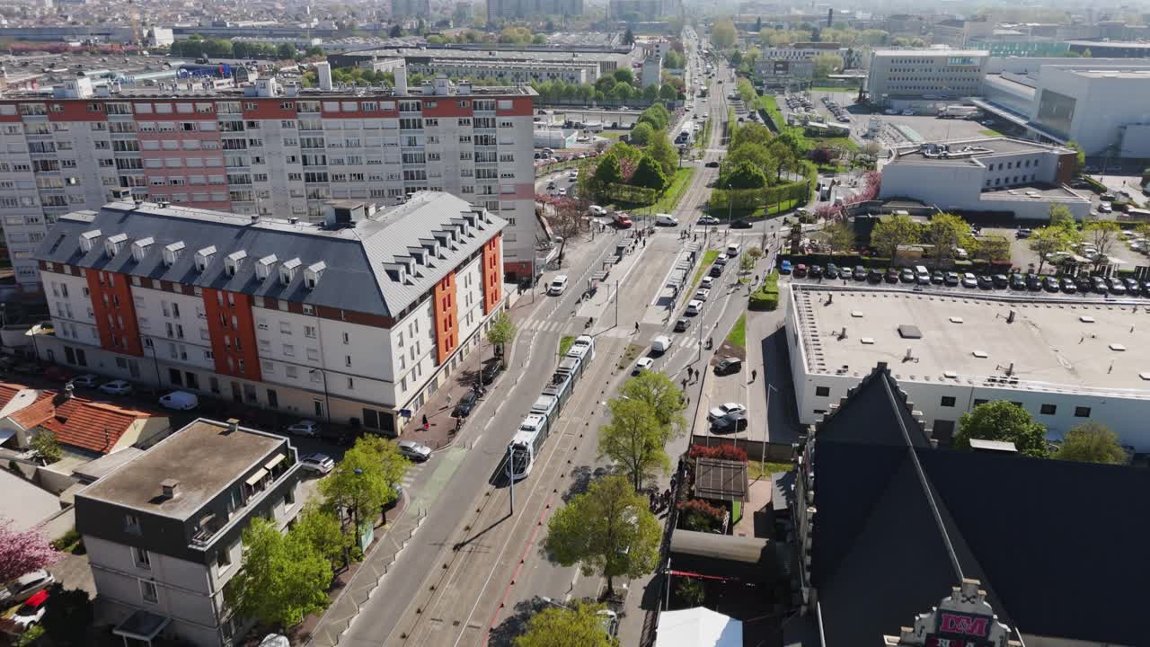 Aerial drone shot tracking a tramway passing near a roundabout, a building, and a shopping mall parking lot in the Paris suburbs - France