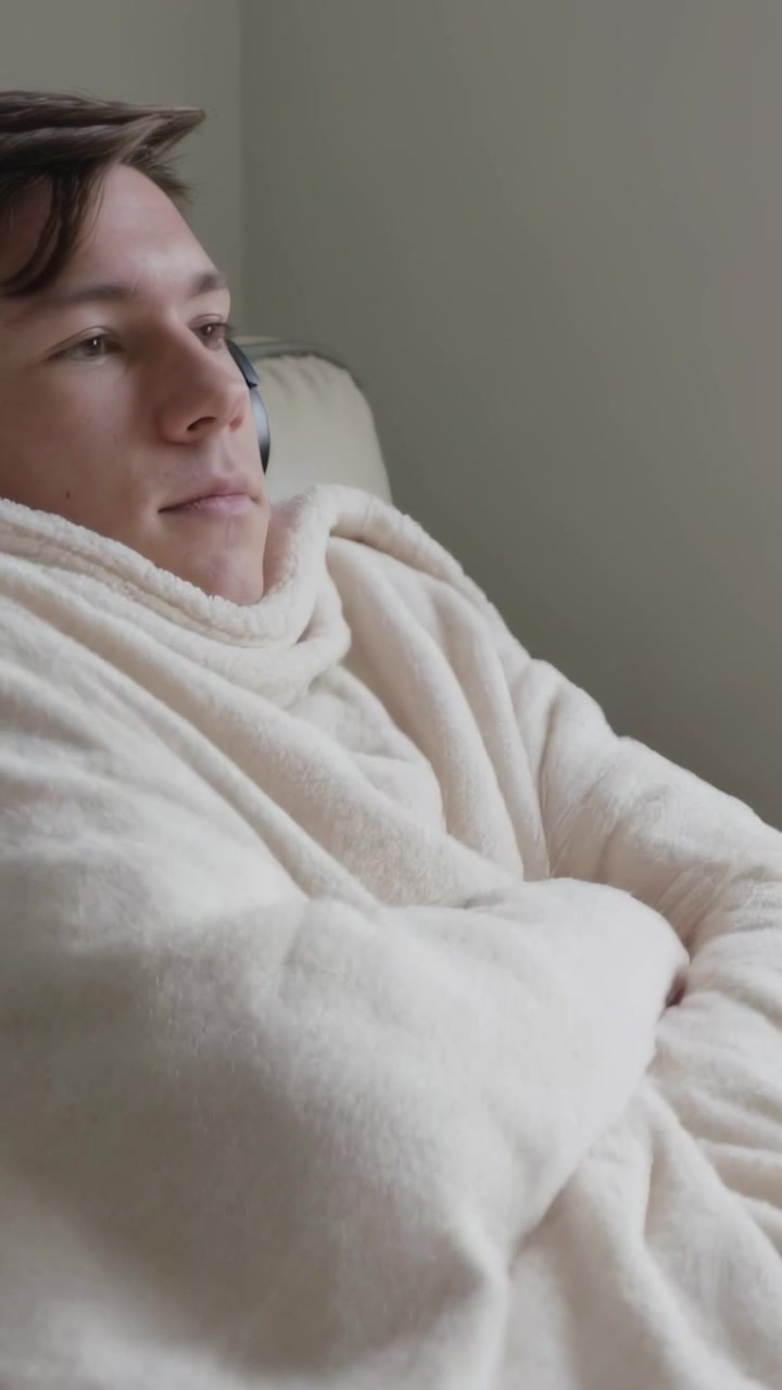 Video verticale: Sentendo freddo, uomo con felpa azzurra che si avvolge in una coperta sul divano con le cuffie