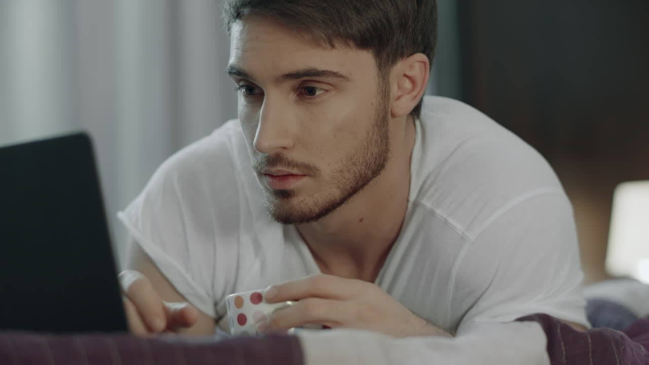 침대에서 커피 컵을 들고 노트북을 사용하는 남자. 남성 블로거가 온라인으로 일하고 있습니다.