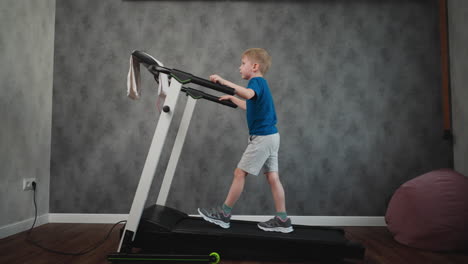 side view of young boy wearing blue shirt and gray shorts walking on treadmill holding handles during home workout session plugged into wall outlet against grey textured wall wooden floor beanbag