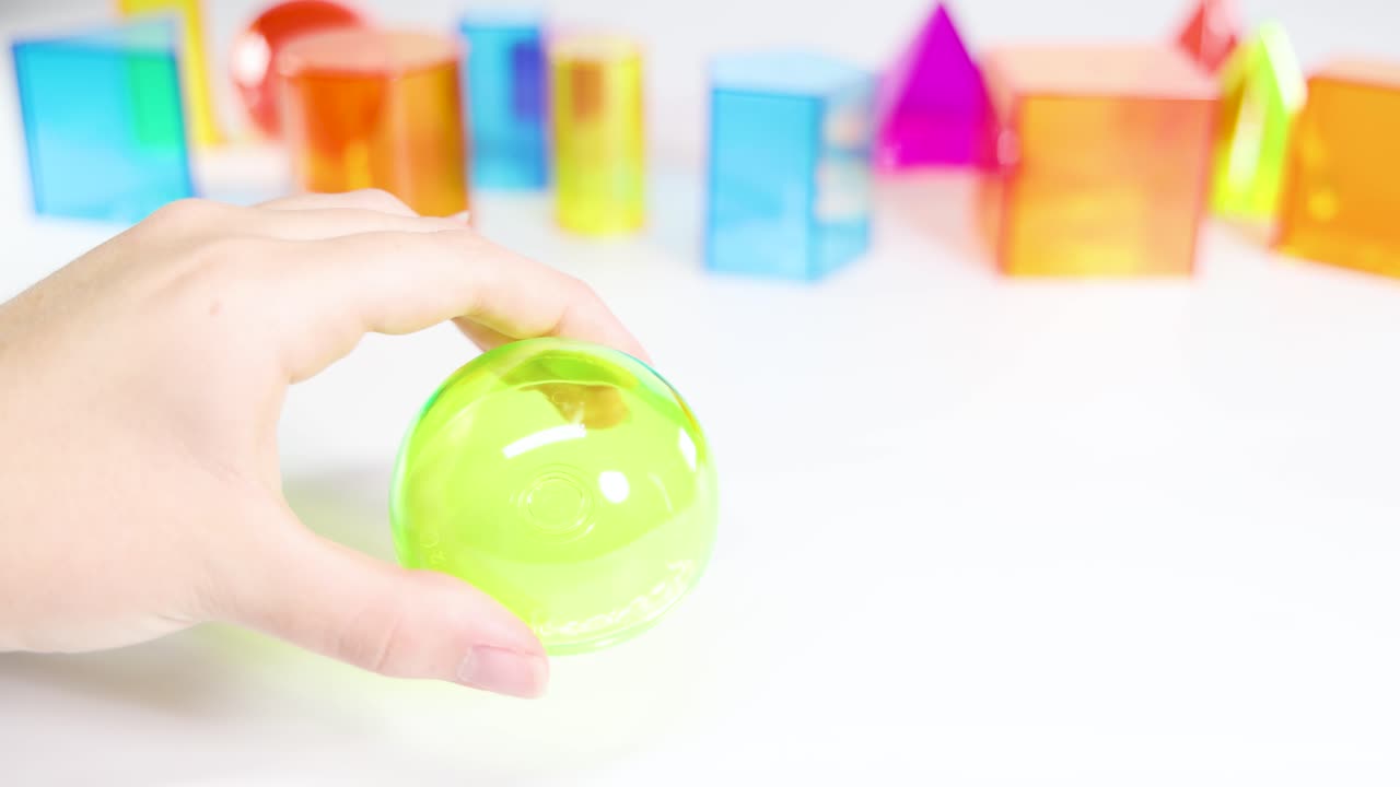 Hands manipulate a translucent green sphere against a backdrop of colorful geometric shapes on a white surface