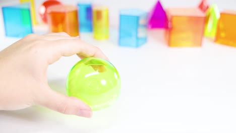 hands manipulate a translucent green sphere against a backdrop of colorful geometric shapes on a white surface