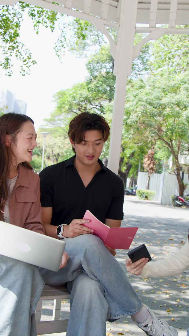 นักเรียนไทยสามคนกำลังทำงานกลุ่มวิชาเรียนร่วมกันโดยใช้แล็ปท็อปและสมุดบันทึกในศาลาที่แสงแดดส่องถึง
