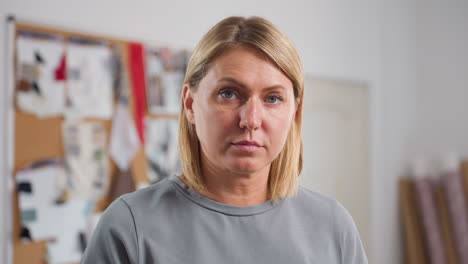 shoulder view of fair skinned woman concentrating deeply on work, head lowered with focused expression, standing inside creative workspace surrounded by blurred inspiration boards and fabric samples