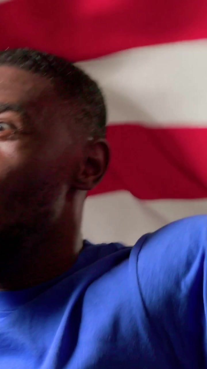 Vídeo vertical: Homem gritando apresentando, puxando a bandeira dos EUA pela lente na parede, com camiseta azul e relógio