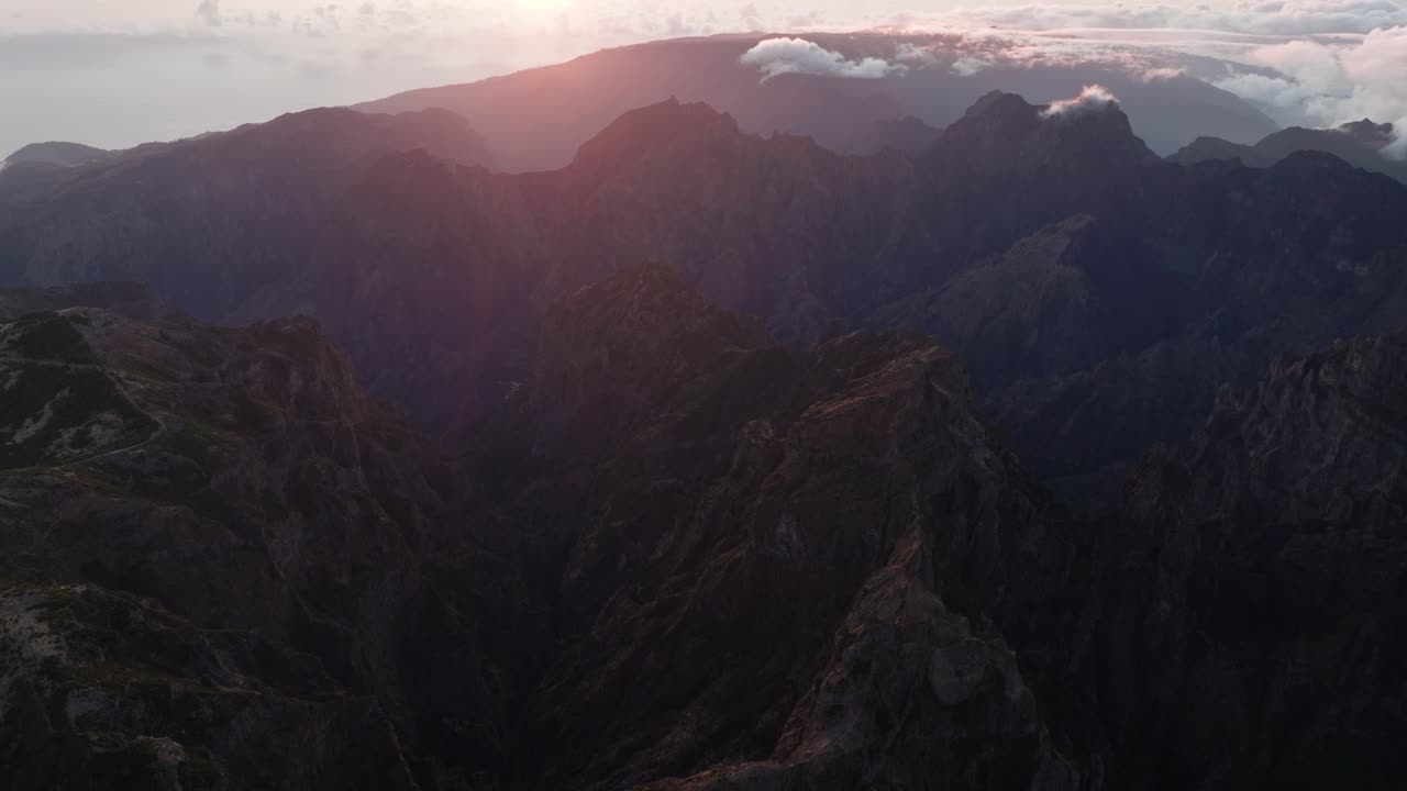 Dramatic aerial of Pico do Arieiro at sunset, casting soft light over jagged cliffs and layered mountain shadows.