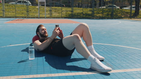 un hombre con pantalones cortos de baloncesto negros y una camisa negra sin mangas está acostado de espaldas en una cancha de baloncestro, sosteniendo su teléfono, con una botella de agua y una pelota de balonceso a su lado.