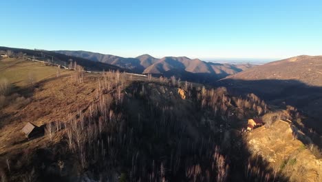 Aerial-view-of-a-hill-top-with-sparse-trees-and-wooden-cabins-in-Transilvania,-Romania