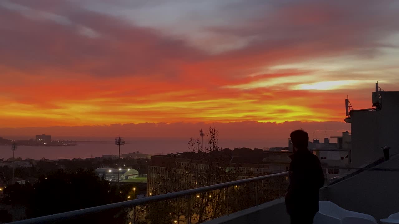 el hombre parado en la terraza mirando el río con la puesta de sol y la costa da caparica al fondo