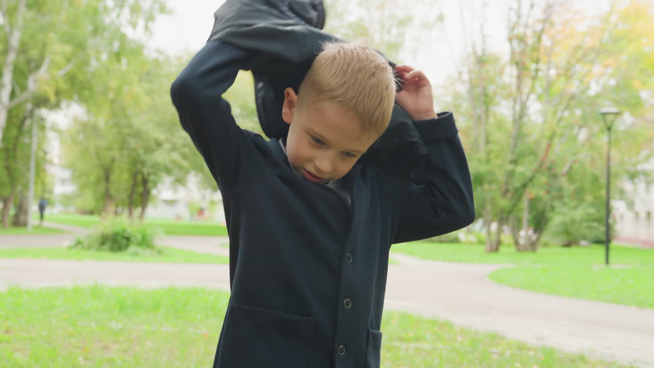 Young white boy putting jacket backtoschool preparation in leafy park smiling while preparing backpack and blazer on cool autumn morning candid preteen student adjusting coat and bag while walking
