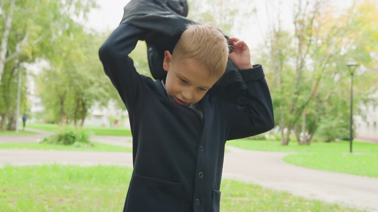 Young white boy putting jacket backtoschool preparation in leafy park smiling while preparing backpack and blazer on cool autumn morning candid preteen student adjusting coat and bag while walking