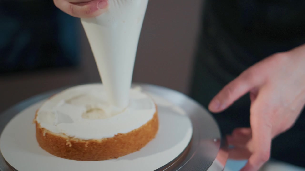 Person carefully pipes white frosting on cake layer. Hands steady as they rotate cake stand. Smooth icing spreads evenly across sponge. Perfect for dessert tutorials or bakery branding
