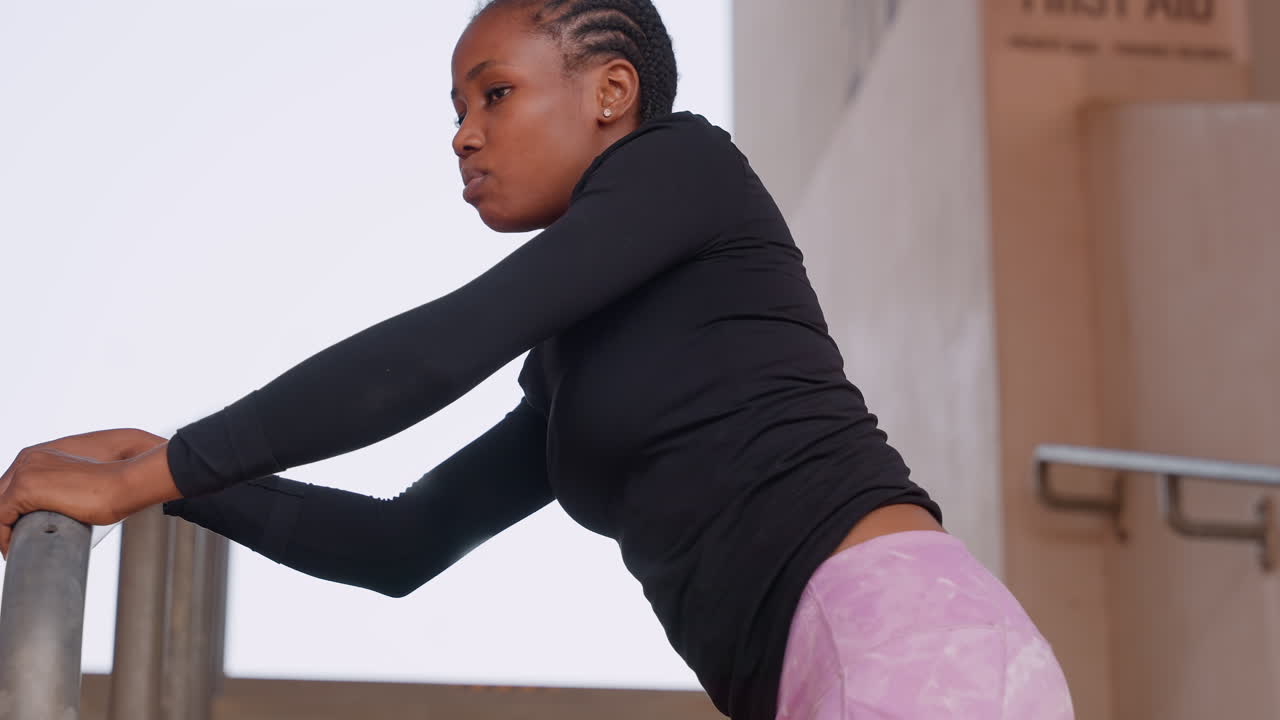 black woman athlete warming on stair railing, focused prerun stretch with braided hair, black long sleeve top and pink leggings, urban concrete stairwell setting, steady breathing and muscle