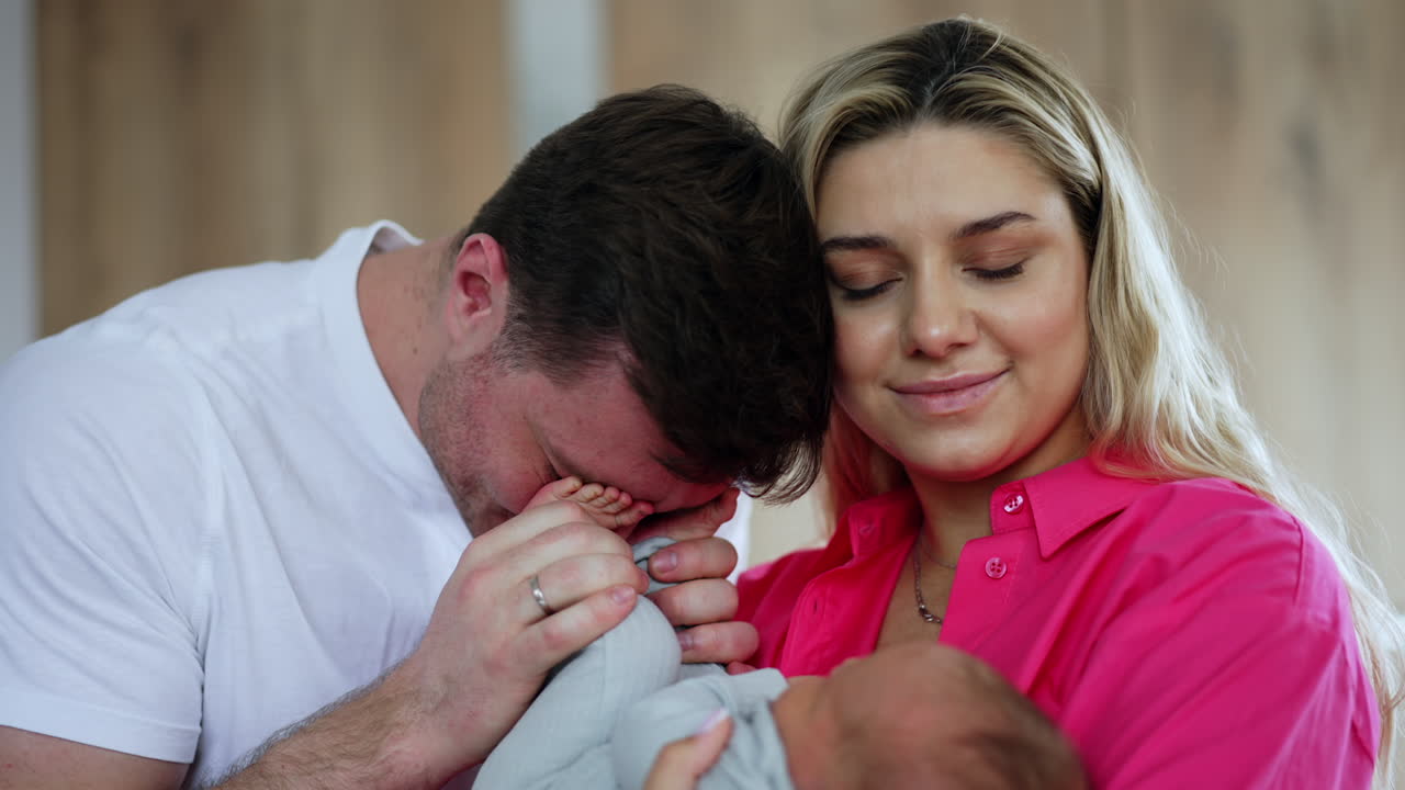 Man and woman with their newborn child. Happy father kisses tiny baby feet and puts them on his eyes. Close up.