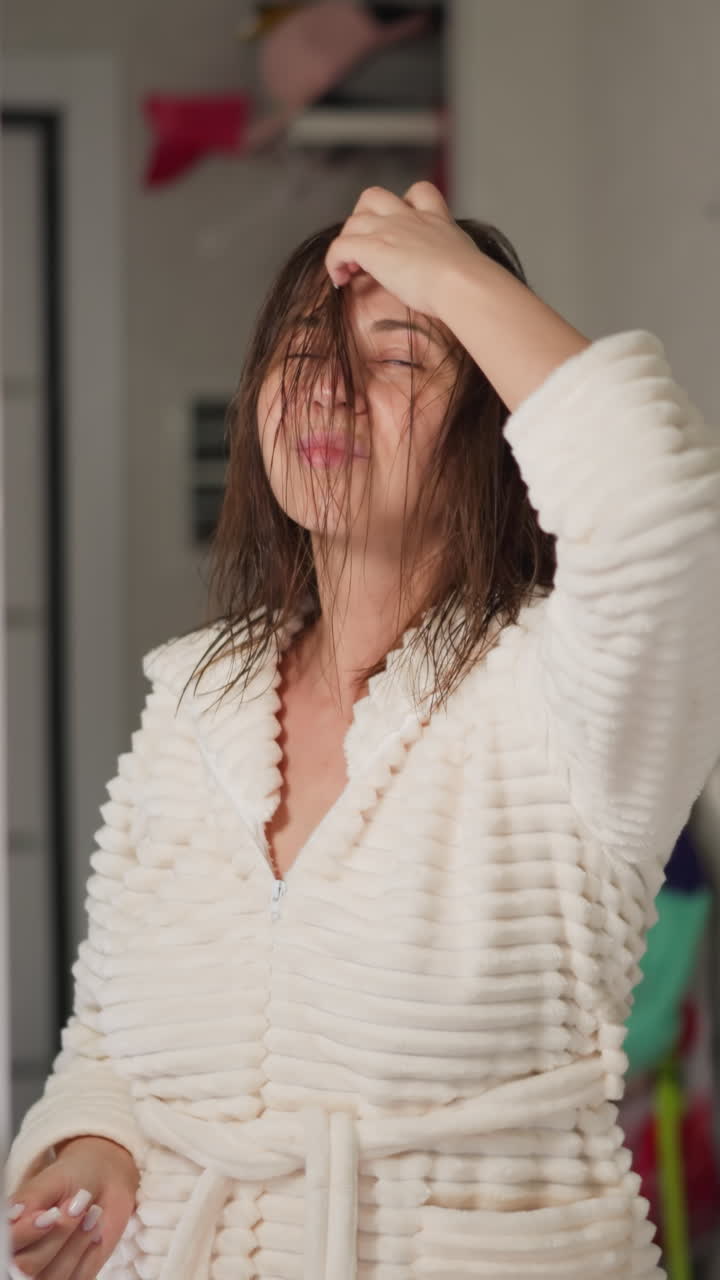 젊은 갈색 여자가 음악을 듣고 샤워를 한 후 은 머리카락을 똑바로 펴고 있습니다. 한 머리카락의 여성은 거울에서 느린 동작을 보고 춤을 니다.