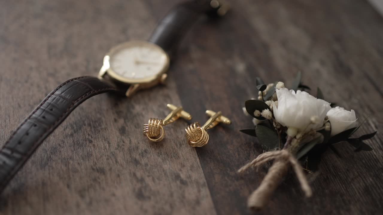 Close up of a gold watch, cufflinks, and a boutonniere arranged on a rustic wooden surface