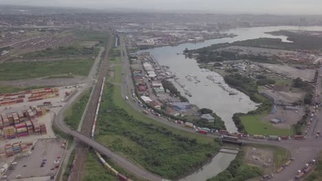 Aerial-footage-of-Durban-harbour-with-yachts-and-traffic-consisting-of-trucks-and-moving-trains-over-a-highway-with-cargo-container-depot-in-view