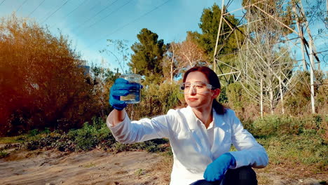 a young woman scientist at a creek, wearing protective eyewear and a lab coat, taking a water sample from the creek, and holding it up to observe it