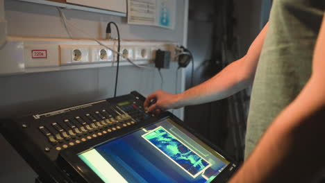 man stands under soft spotlight in dim technical room facing workstation near corkboard filled with handwritten notes and charts, while electronic panels and glowing blue lights are visible in background