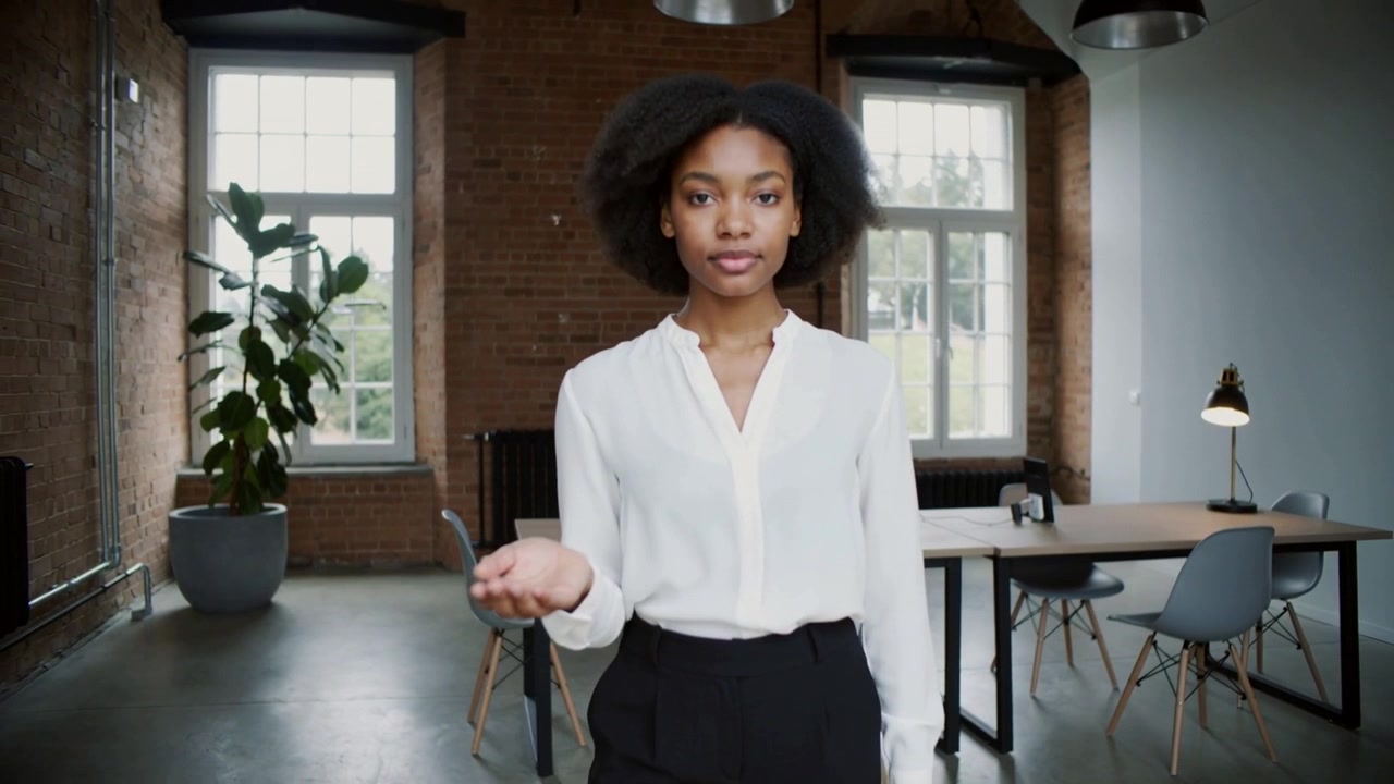 Walking from left woman in white blouse turning to camera at loft, presenting palm over desk