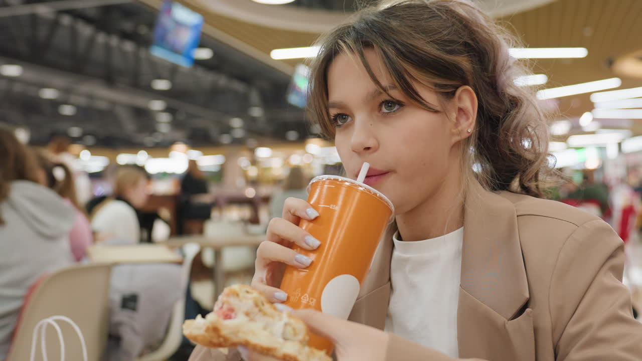 Empresario caucásico comiendo pizza en la hora punta del almuerzo, vaso naranja a un lado, consultando el teléfono entre bocado y bocado, expresión concentrada, chaqueta informal, ajetreo en la zona de restauración, momento de tentempié multitarea