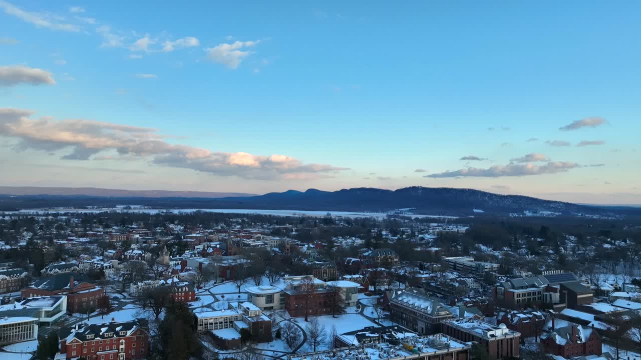 Drone footage of Northampton at twilight, highlighting snow-covered rooftops, historic architecture, and the serene backdrop of distant mountains under a clear winter sky