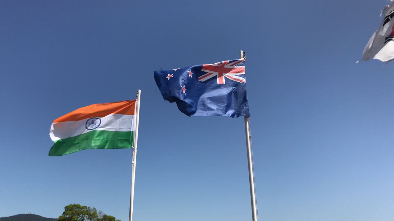 The national flags of India and New Zealand flutter in the wind under a clear blue sky with part of the Korean flag seen waving in the left upper corner