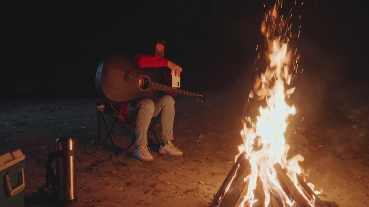 Sessão noturna de violão na fogueira. Campista solo masculino asiático dedilha um violão acústico em uma cadeira dobrável ao lado de uma fogueira crepitante, sob a luz quente do fogo, com caixa térmica e garrafa térmica por perto, em um clima reflexivo e com um dedilhado suave.