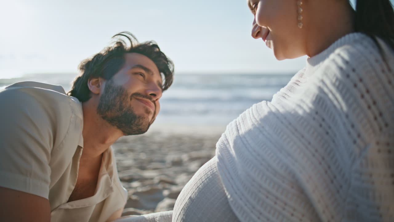사랑하는 남편이 배에 키스하는 임신한 아내가 해가 지는 바가에 앉아 있습니다.