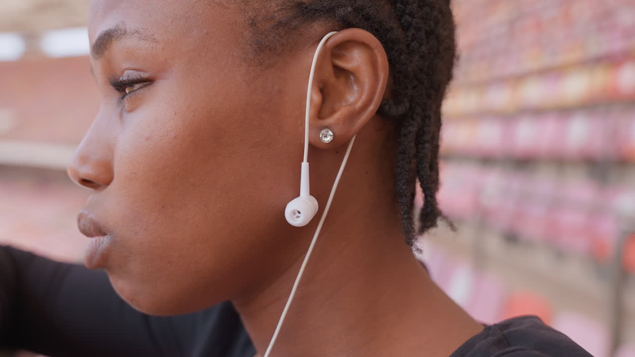 Primo piano di una donna con auricolari in un momento contemplativo pre-partita. Profilo laterale con capelli intrecciati, una sottile traccia di sudore sulla guancia e sedili sfocati dello stadio sullo sfondo. Luce ambientale e un'espressione concentrata.