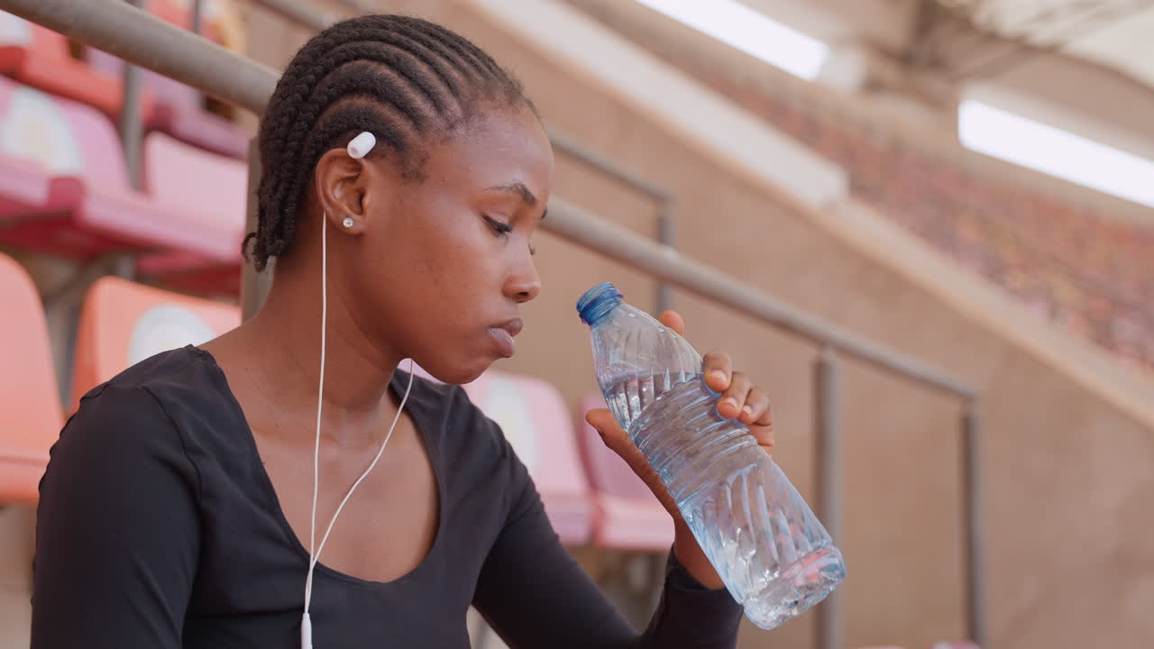 Teen girl drinking water. Young woman with braided hair sips water during rest period. Teenage girl with braided hairstyle and earphones drinks from plastic bottle during training break