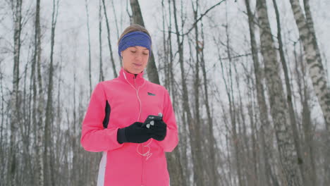 Una-Joven-Que-Hace-Jogging-Por-La-Mañana-Sostiene-En-Sus-Manos-Un-Teléfono-Inteligente-Que-Elige-Una-Pista-De-Música-Para-Entrenar.-Carrera-De-Invierno.-Utilice-Dispositivos-Y-Aplicaciones-De-Formación-Modernos.