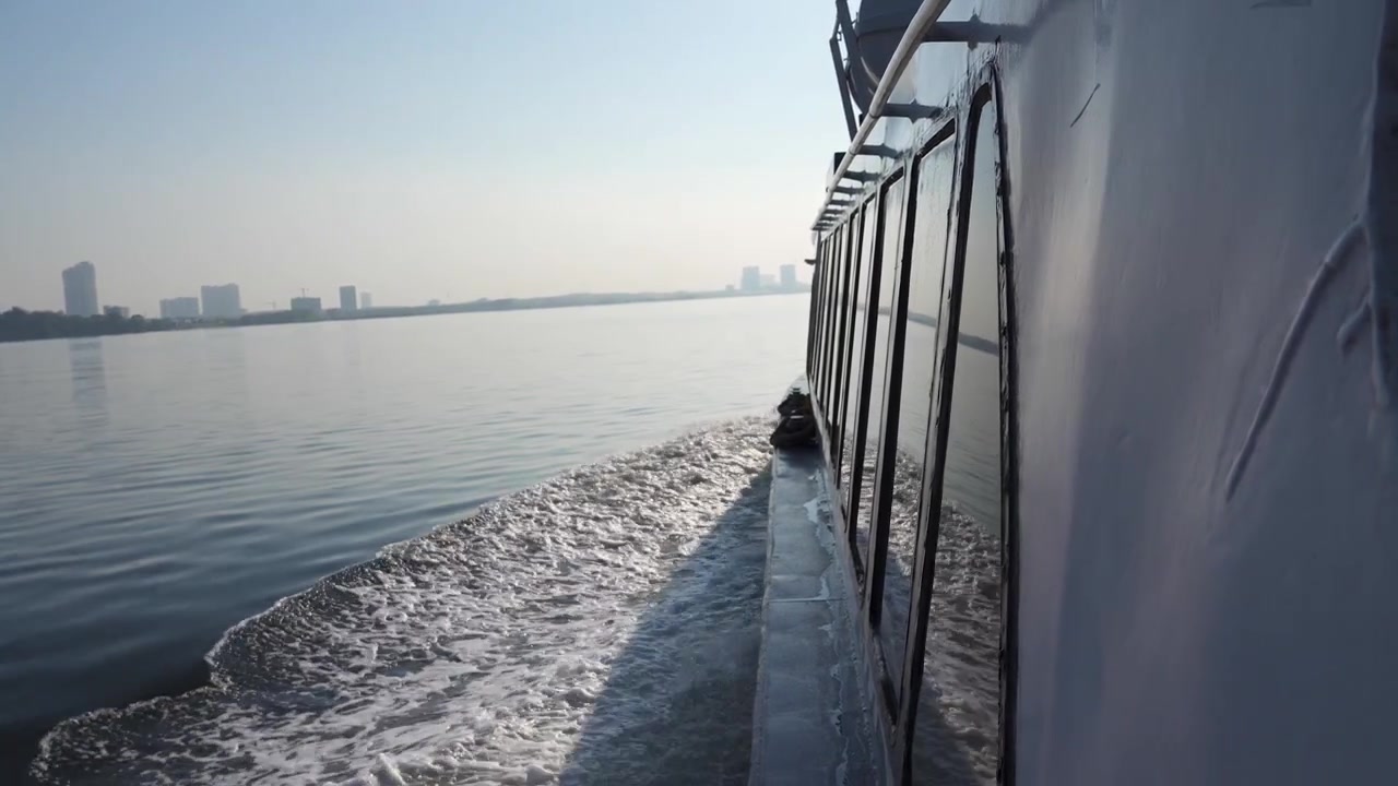 Eine Aufnahme aus der Ich-Perspektive vom Deck einer fahrenden Fähre, die eine weiße Schaumspur und eine entfernte Stadtsilhouette entlang einer wichtigen Schifffahrtsroute in Südostasien oder der Straße von Malakka zeigt. Reise- und maritime Themen.