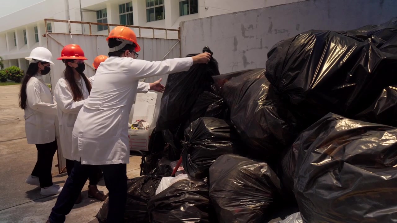Workers in lab coats, safety helmets, and face masks diligently sort and move plastic waste bags at a recycling plant in Mexico City, contributing to eco-friendly construction materials production.