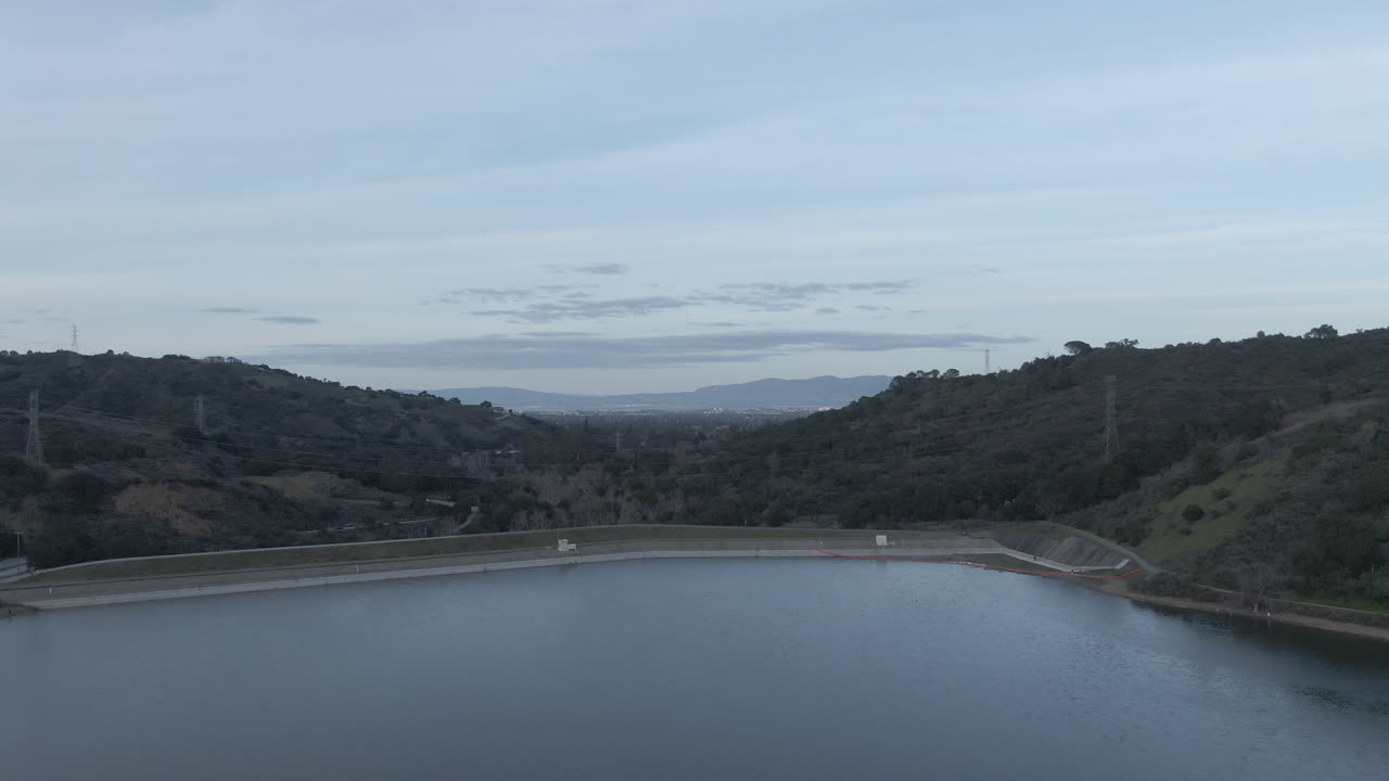 A cinematic wide aerial shot overlooking the calm waters of Steven Creek Reservoir nestled in the Santa Cruz Mountains. The footage captures the surrounding green hills and the concrete dam structure
